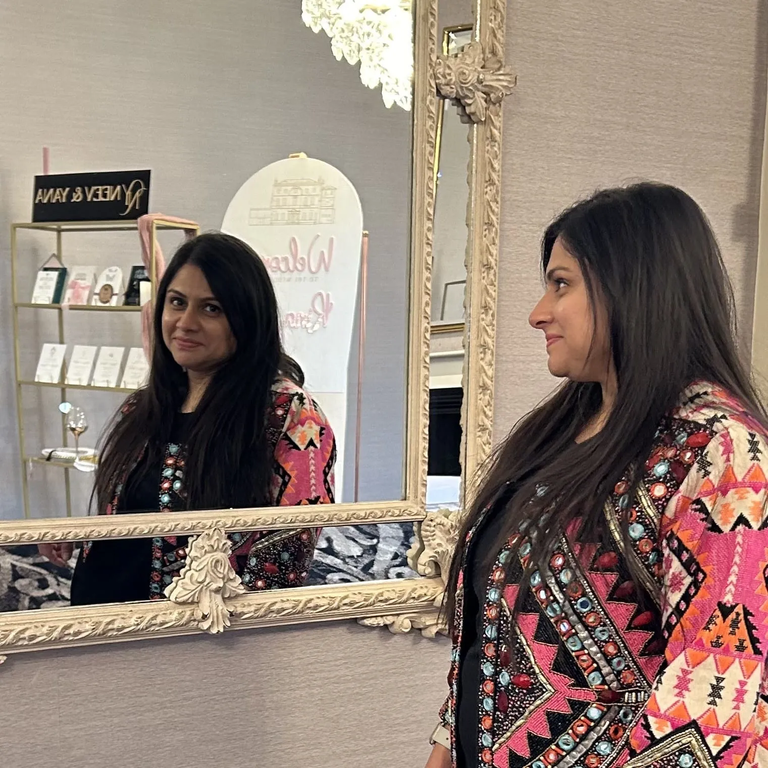 Neh at a wedding show stand, looking into an ornate mirror, with the Neev and Yana sign and a shelf of stationery samples behind her.