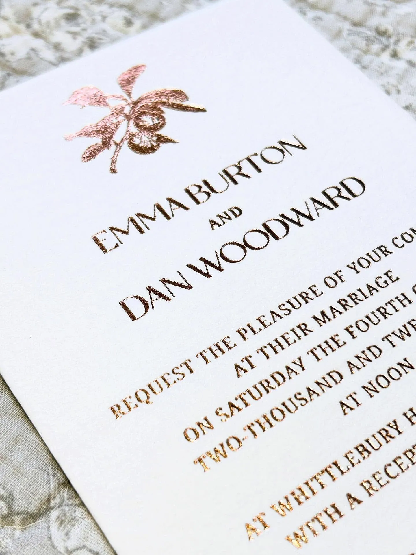 Oblique close-up of a wedding invitation in copper hot foil on ivory cotton, lying on grey lace, with a foiled botanical sprig above the names.