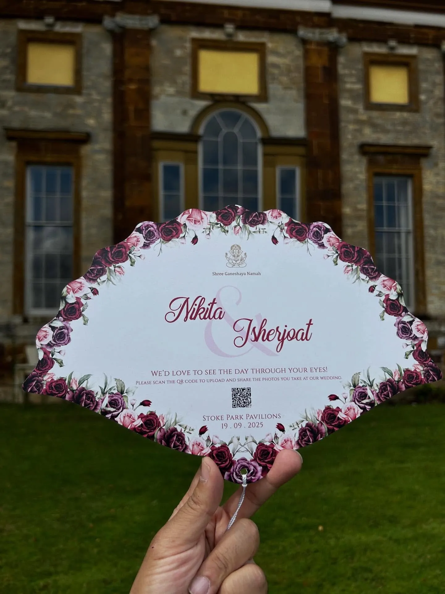 Fan-shaped wedding keepsake card with a painted rose border and a QR code, held up in front of a country house.