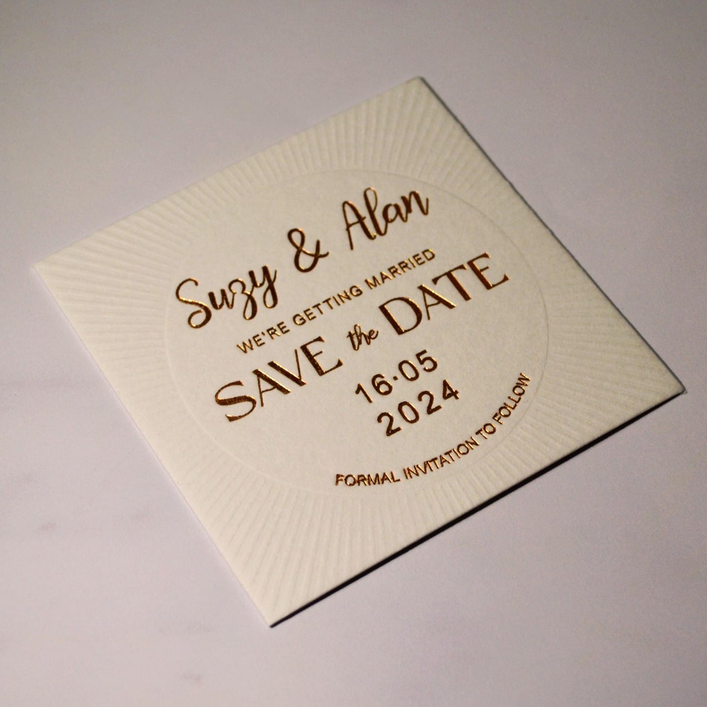 Square ivory save-the-date with sunburst debossing radiating from the centre, lettered in gold hot foil, photographed at an angle in raking light so the impression is visible.