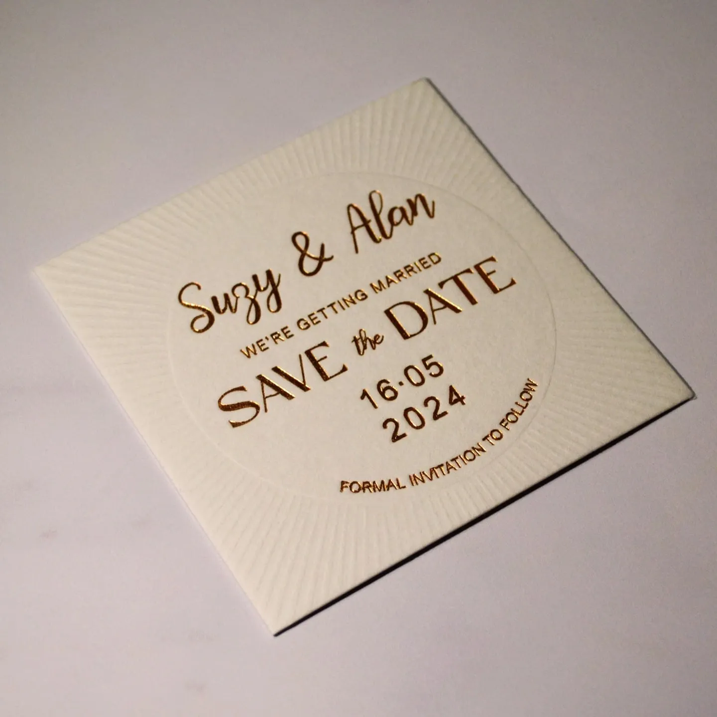 Square ivory save-the-date with sunburst debossing radiating from the centre, lettered in gold hot foil, photographed at an angle in raking light so the impression is visible.