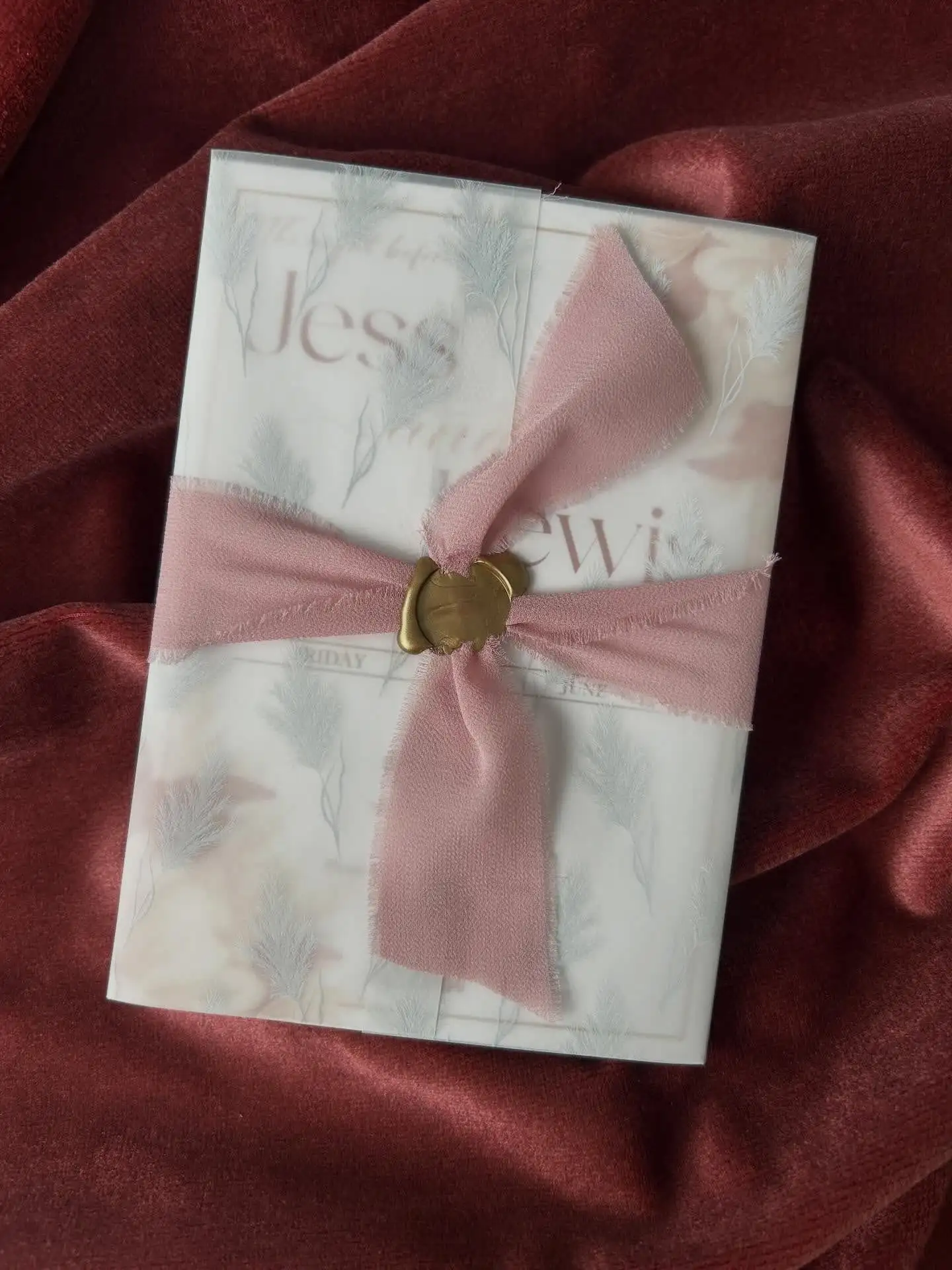 Wedding invitation suite wrapped in printed vellum, tied with frayed blush silk ribbon and a gold wax seal, photographed on deep red velvet.
