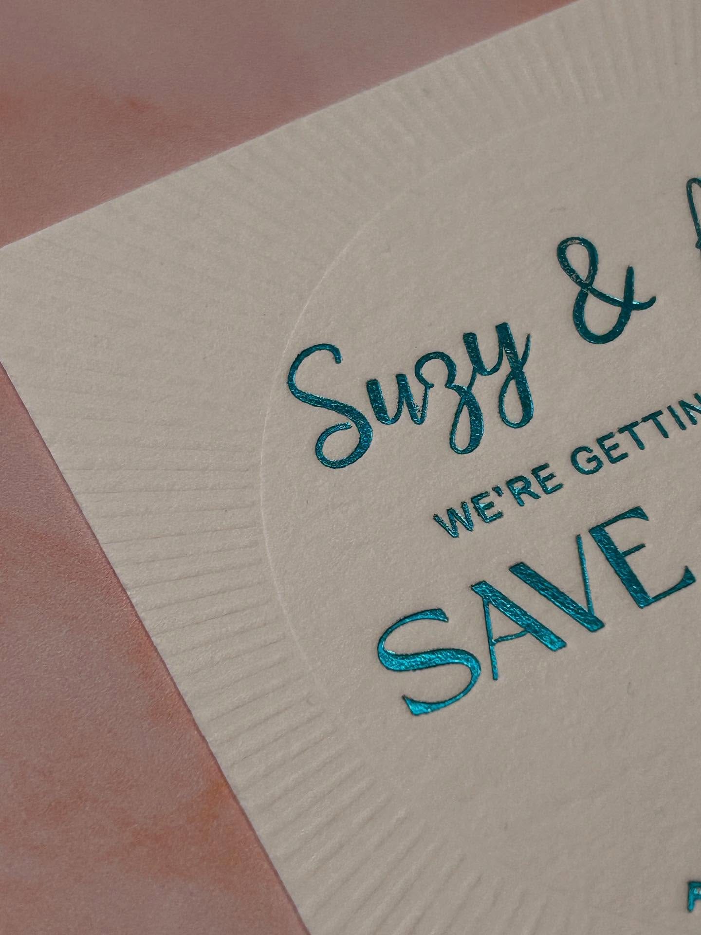 Close-up of an ivory save-the-date at an angle, with teal hot foil script catching the light and blind-debossed sunburst rays behind it.