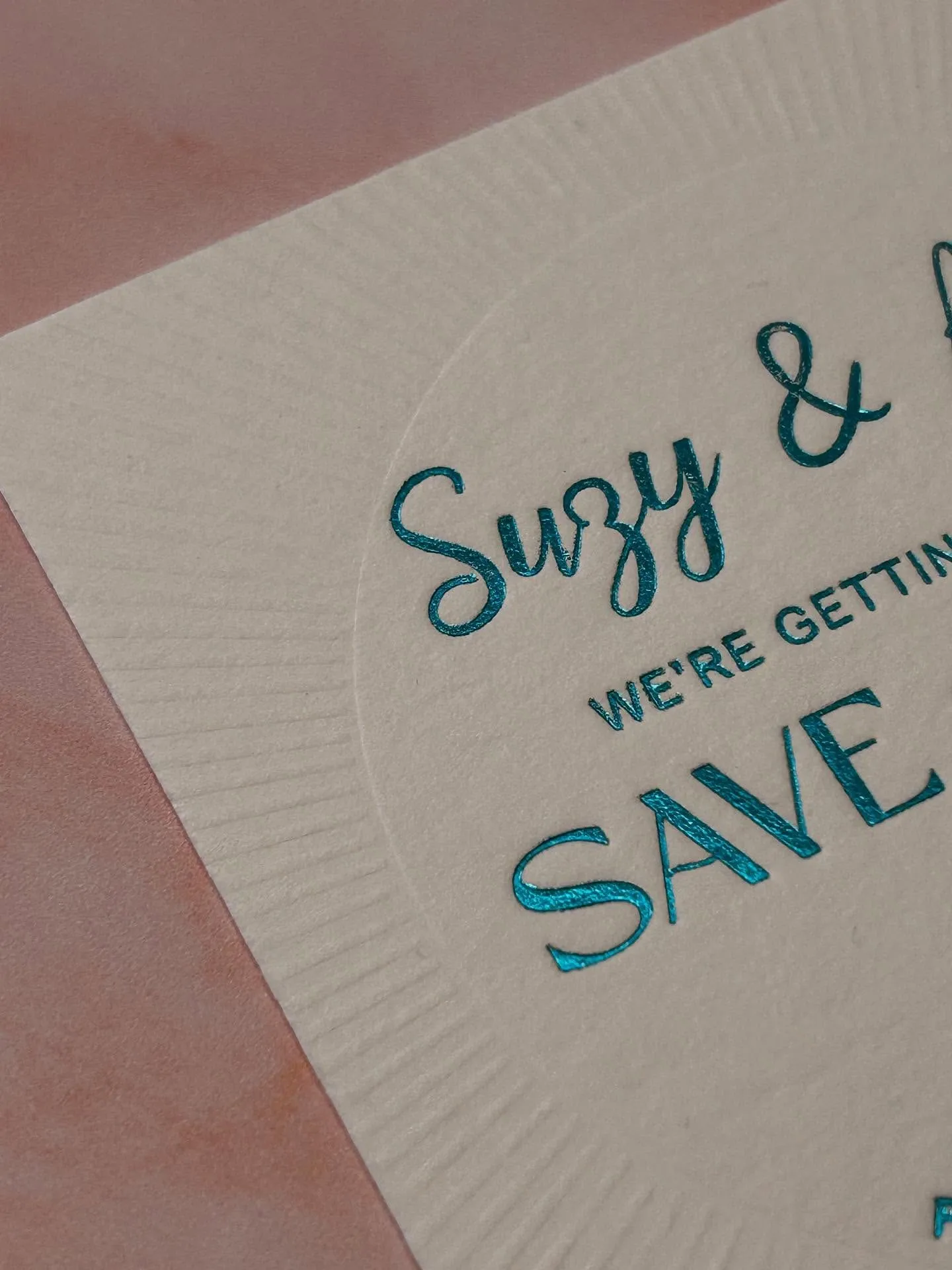 Close-up of an ivory save-the-date at an angle, with teal hot foil script catching the light and blind-debossed sunburst rays behind it.