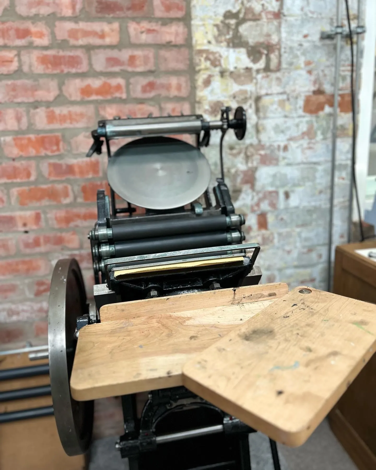 The studio's Gem No1 platen letterpress against exposed brick, showing the ink disc, rollers, flywheel and two wooden feed boards.